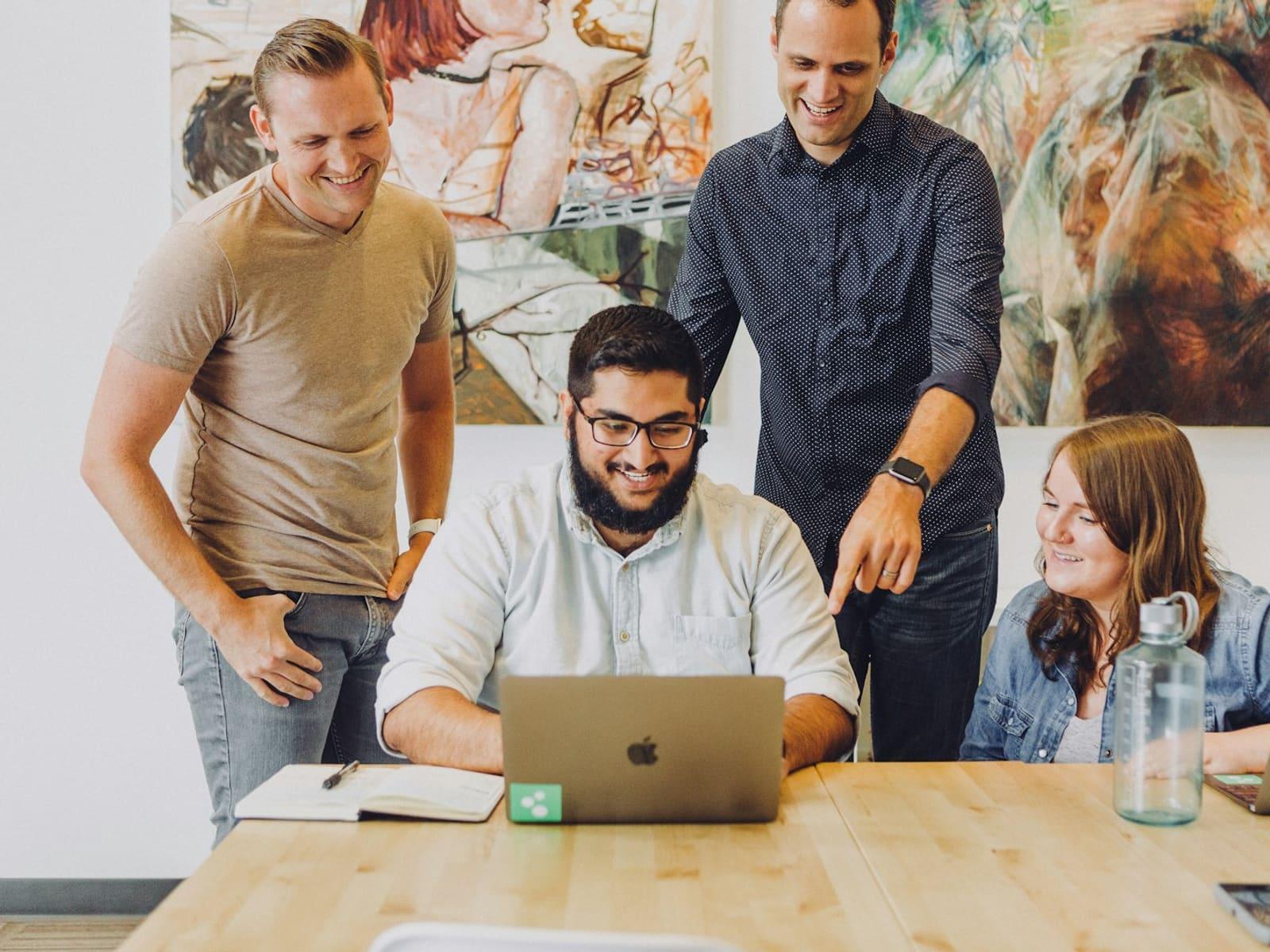 Four coworkers smiling together around a laptop at a wooden table